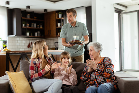 Heartwarming scene unfolds as a multi-generational family gathers on a couch to present a birthday cake to a delighted grandmother, creating memories to cherishの写真素材