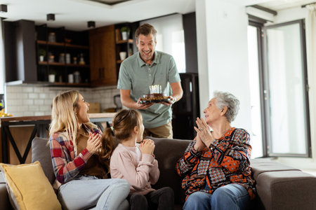 Heartwarming scene unfolds as a multi-generational family gathers on a couch to present a birthday cake to a delighted grandmother, creating memories to cherishの写真素材