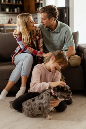 Joyful parents watch their daughter play with a happy french bulldog at homeの写真素材