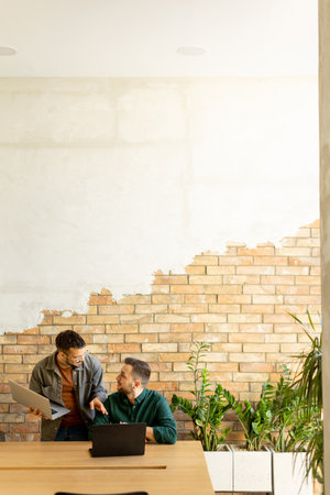 Two smiling professionals engage in a collaborative work session at a wooden table, their camaraderie evident in a contemporary office setting with an exposed brick wall backdropの写真素材