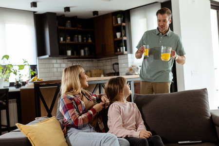 A smiling father brings a tray of orange juice to his happy family seated in a living roomの写真素材