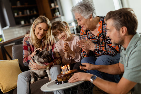 Heartwarming scene unfolds as a multi-generational family gathers on a couch to present a birthday cake to a delighted grandmother, creating memories to cherishの写真素材