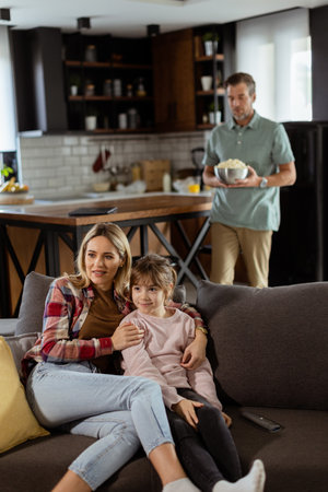 A family of three is comfortably nestled on a couch, their faces reflecting excitement and attentiveness as they share a bowl of popcorn during a suspenseful movie nightの写真素材