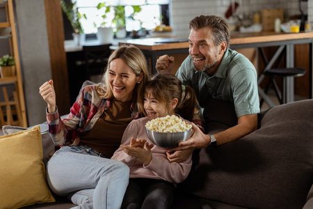 A family of three is comfortably nestled on a couch, their faces reflecting excitement and attentiveness as they share a bowl of popcorn during a suspenseful movie nightの写真素材