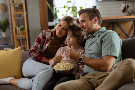 A family of three is comfortably nestled on a couch, their faces reflecting excitement and attentiveness as they share a bowl of popcorn during a suspenseful movie nightの写真素材