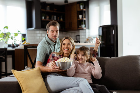 A family of three is comfortably nestled on a couch, their faces reflecting excitement and attentiveness as they share a bowl of popcorn during a suspenseful movie nightの写真素材
