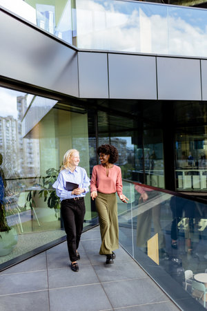 Two colleagues laugh together, one holding a tablet in a sunny, glass-walled workspace.の写真素材