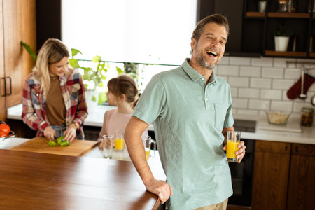 Smiling man holds a glass of juice while two women cook in a bright kitchenの写真素材