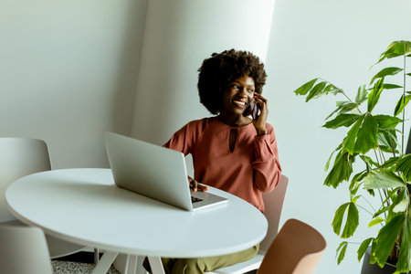 A cheerful woman talks on her phone at a cafe table with a laptop and a vibrant plant beside her.の写真素材