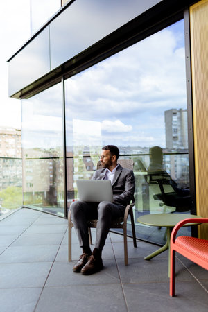 A focused man in a suit uses his laptop on a sunny balcony overlooking the cityscape.の写真素材
