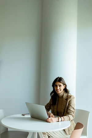 Focused businesswoman in a stylish blazer types away on her laptop at a sleek white table, surrounded by minimalist decor.の写真素材