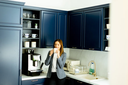 A smiling woman in business attire holding a coffee mug, taking a relaxing break in a stylish blue kitchen.の写真素材