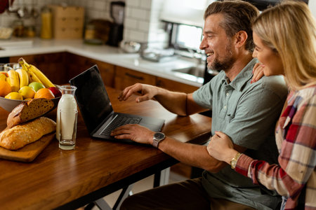 A cheerful couple enjoys a light-hearted moment in their sunny kitchen, working on laptop surrounded by a healthy breakfastの写真素材