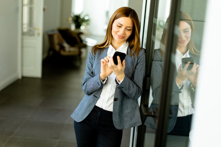 A cheerful professional woman stands in an office corridor, her content reflection visible as she clutches a smartphone.の写真素材