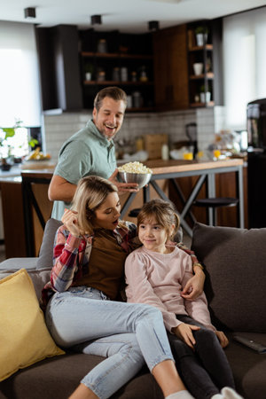 A family of three is comfortably nestled on a couch, their faces reflecting excitement and attentiveness as they share a bowl of popcorn during a suspenseful movie nightの写真素材