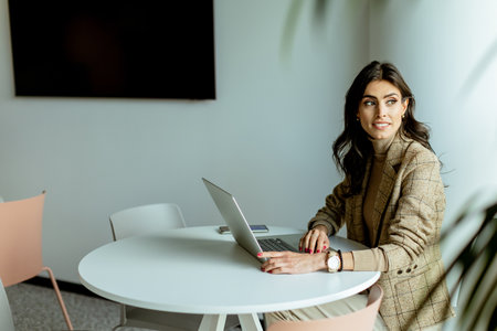 Focused businesswoman in a stylish blazer types away on her laptop at a sleek white table, surrounded by minimalist decor.の写真素材