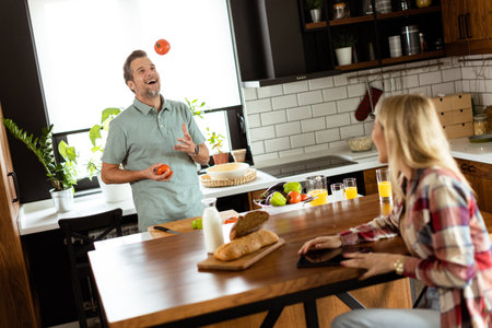 Woman reads from a tablet at the kitchen counter while a man holding an tomatoes smiles at herの写真素材