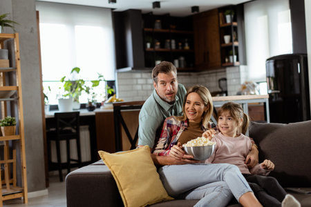 A family of three is comfortably nestled on a couch, their faces reflecting excitement and attentiveness as they share a bowl of popcorn during a suspenseful movie nightの写真素材