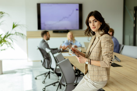 A poised woman with a tablet stands at the forefront of a collaborative office space, her team engaged in discussion behind her.の写真素材
