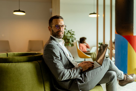 Focused businessman works on his laptop while sitting on a plush green couch in a vibrant, contemporary workspace.の写真素材