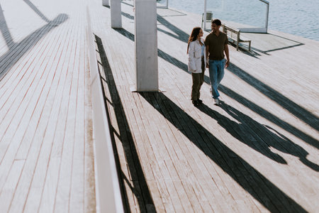 Young couple walks hand in hand, casting long shadows on the wooden boardwalk by the sparkling sea in Barcelonaの写真素材