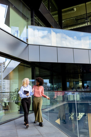 Two colleagues laugh together, one holding a tablet in a sunny, glass-walled workspace.の写真素材