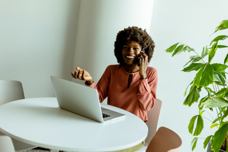 Confident woman multitasks with a phone call and laptop in a bright, plant-adorned office, conveying casual professionalism.の写真素材