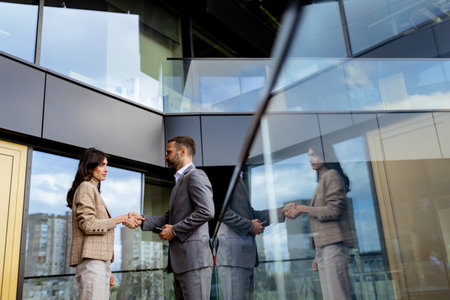 Two business professionals shake hands outside a contemporary building, their interaction mirrored in the glass.の写真素材