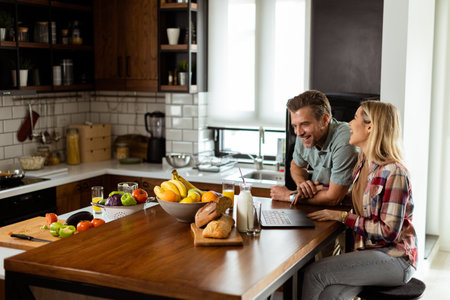 A cheerful couple enjoys a light-hearted moment in their sunny kitchen, working on laptop surrounded by a healthy breakfastの写真素材