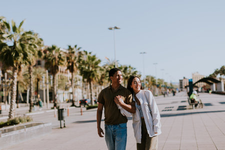 Young couple holds hands and smiles as they walk down a sunlit path in Barcelona, framed by palmsの写真素材