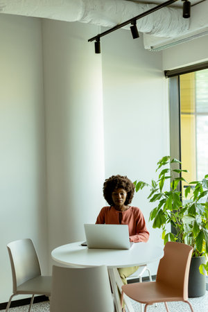 A woman focused on her laptop at a bright cafÃ© table by a large window next to indoor plants.の写真素材