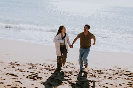 A smiling couple enjoys a carefree run on a sandy shore, waves gently breaking in the backgroundの写真素材
