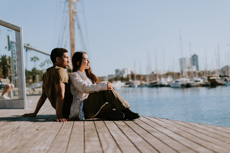 Couple sits closely on a dock, enjoying a peaceful moment overlooking the marina on a sunny day in Barcelona.の写真素材