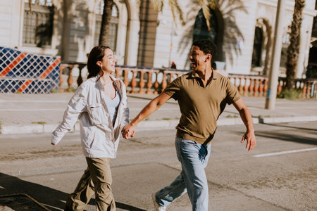 Young couple holds hands and smiles as they walk down a sunlit path in Barcelona, framed by palmsの写真素材
