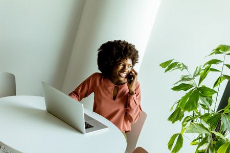 A cheerful woman talks on her phone at a cafe table with a laptop and a vibrant plant beside her.の写真素材