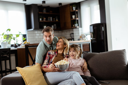A family of three is comfortably nestled on a couch, their faces reflecting excitement and attentiveness as they share a bowl of popcorn during a suspenseful movie nightの写真素材
