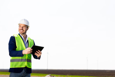 A safety-helmeted engineer with clipboard scrutinizes the workings of a majestic wind turbine amidst lush greenery under a clear sky.の写真素材
