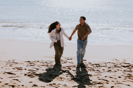 A smiling couple enjoys a carefree run on a sandy shore, waves gently breaking in the backgroundの写真素材