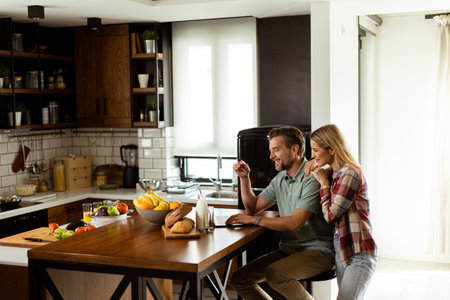 A cheerful couple enjoys a light-hearted moment in their sunny kitchen, working on laptop surrounded by a healthy breakfastの写真素材