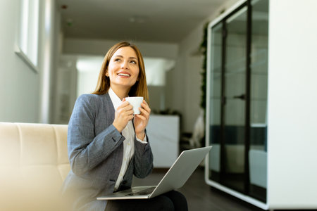 Poised professional sips her coffee while engaging with her laptop, basking in soft daylight.の写真素材