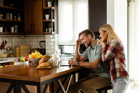 A cheerful couple enjoys a light-hearted moment in their sunny kitchen, working on laptop surrounded by a healthy breakfastの写真素材
