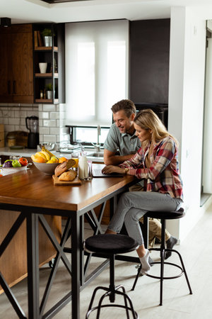 A cheerful couple enjoys a light-hearted moment in their sunny kitchen, working on laptop surrounded by a healthy breakfastの写真素材
