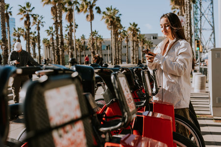 Young woman stands by a row of red bikes in sunny Barcelona, checking her phone, likely using a bike-sharing appの写真素材
