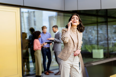 A stylish woman converses on her smartphone, walking past colleagues gathered in casual conversationの写真素材