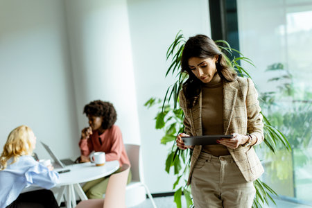 Stylishly dressed woman with a tablet stands in a modern office, her colleagues engaged in work behind herの写真素材