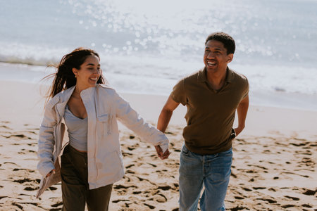 A smiling couple enjoys a carefree run on a sandy shore, waves gently breaking in the backgroundの写真素材