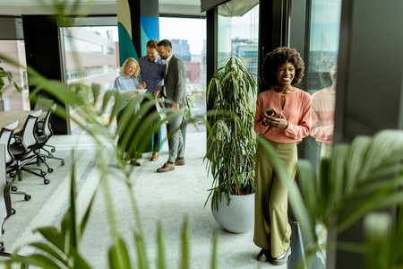African American woman on her phone stands by lush indoor plants, while colleagues converse in the backgroundの写真素材
