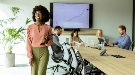 Confident African American woman stands with a tablet in a meeting room, colleagues blurred in the background.の写真素材