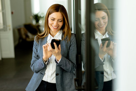 A cheerful professional woman stands in an office corridor, her content reflection visible as she clutches a smartphone.の写真素材