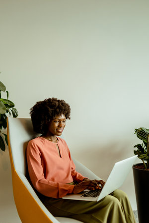 Spirited African American businesswoman smiling while working on her laptop, comfortably seated in a vibrant office lounge.の写真素材